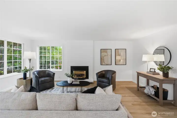 Sun-drenched living room anchored by a white brick fireplace with brass insert, oversized windows overlooking the greenery, and warm wood-look floors.
