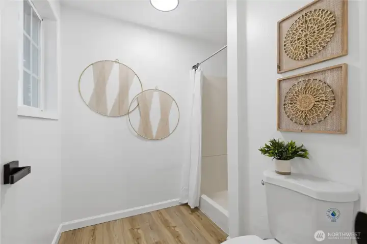 Shower and water closet area with fresh finishes, wood-look flooring, and a natural window for ventilation and light.