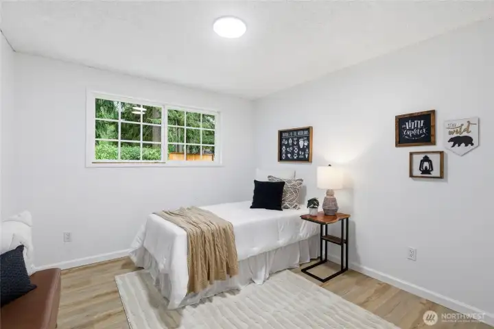 Cozy bedroom with a wide window overlooking the backyard greenery and fresh wood-look flooring throughout.