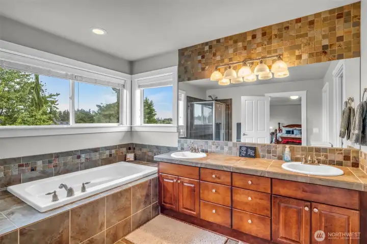 Luxurious 5-piece primary bathroom showcasing dual vanities, extensive cabinetry, tile finishes, a soaking tub, and large windows that fill the space with natural light. Luxurious 5-piece primary bathroom showcasing dual vanities, extensive cabinetry, tile finishes, a soaking tub, and large windows that fill the space with natural light.