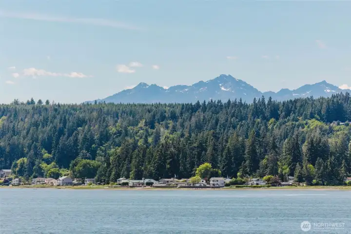 No enhancement. No filter. This is what the Hood Canal looks like on a clear Pacific Northwest day. The snow-capped Olympic Mountain peaks rise dramatically above a dense wall of ancient evergreen forest, their jagged silhouettes painted against a brilliant blue sky. The canal stretches wide and calm in the foreground, its blue-green water reflecting the light in a way that changes hour by hour, season by season, and never gets old.  A quiet, charming waterfront community nestles along the near shore — modest, unpretentious, and utterly at peace with its surroundings. This is not a view from a vacation rental or a hotel balcony. This is the view from home. Your home.  There are people who drive hours to see scenery like this on a weekend trip. There are people who save for years to vacation somewhere that looks like this. And then there are the lucky few who simply wake up here every day because they had the wisdom to make this place their own.