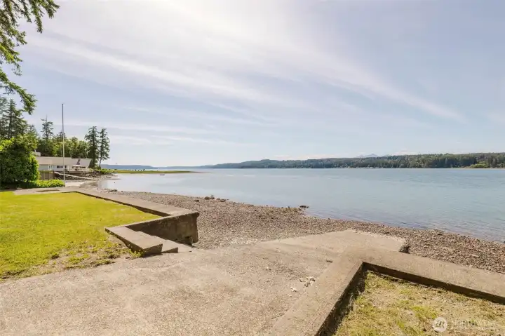 This is the view that seals the deal. Standing at the water's edge on your own property, Hood Canal stretches wide and brilliant in both directions — the rocky shoreline curving away to the left, the forested hills of the far shore reflected in the shimmering water, and mountains rising softly on the horizon under a vast Pacific Northwest sky. This is yours. Every single day.  The concrete boat ramp sits right here on the property — and while it is not configured for launching directly from the property, that is hardly a limitation on the water. Public boat launches are just a short distance away, meaning you can launch your boat, cruise right over, and tie up at your own waterfront. Come and go from the water on your own terms, on your own schedule, from your own beach.  A lush green lawn runs right to the bulkhead edge — perfect for lawn chairs, a fire pit, morning stretches, or simply standing here and letting the view remind you why you made the best decision of your life.