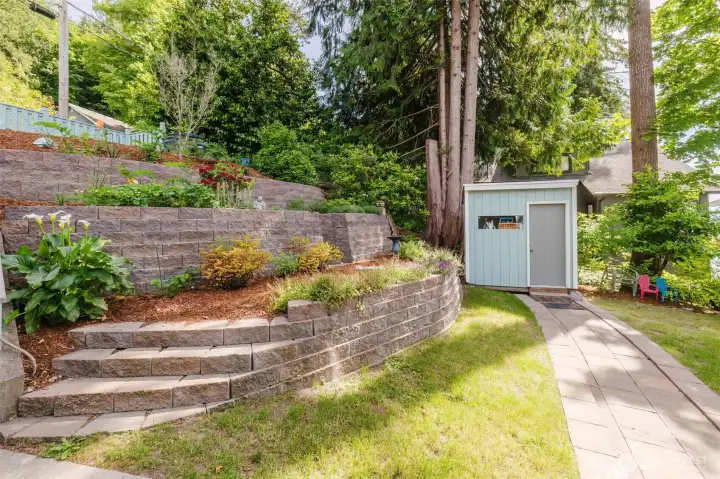 A garden that stops people in their tracks.  This isn't just a yard — it's a $42,000 outdoor sanctuary crafted with intention. Sweeping multi-tiered retaining walls built from rich, textured stone create dramatic level changes that feel both grand and intimate at the same time. Each terrace has been thoughtfully planted with a curated mix of flowering plants, lush shrubs, ornamental grasses, and established perennials — calla lilies, roses, lavender, and more burst with color across every level.Towering Pacific Northwest evergreens and mature deciduous trees frame the space, casting dappled shade and giving the entire property a sense of privacy and seclusion that is almost impossible to find on a waterfront lot. Meandering stone steps guide you through each level of the garden, past birdbaths and garden art that give the space a warm, personal character. A freshly painted storage shed matches the home's exterior perfectly — practical without sacrificing a single ounce of charm.