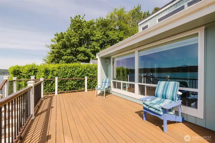 Fifty thousand dollars of pure outdoor living, and every penny of it shows. This sweeping waterfront deck stretches generously across the entire back of the home — wide enough for dining, lounging, entertaining, and simply doing nothing at all while Hood Canal glitters in front of you and a mountain peak sits perfectly framed on the horizon.  The rich warm decking boards glow in the afternoon sun, and the classic white and dark rail system gives the whole space a crisp, finished look that feels both elevated and completely at ease. A lush mature hedge along one side creates a natural privacy screen without blocking a single inch of that breathtaking water view straight ahead.  The large sliding glass windows of the living room and dining room open directly onto the deck, blurring the line between inside and out in the most effortless way. On a warm Pacific Northwest evening, slide them open, set out a few chairs, and let the outside in completely.