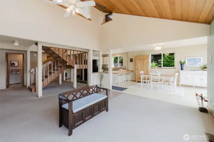Soaring vaulted ceilings with warm wood planking draw your eye up and give the entire main floor a sense of grandeur that feels anything but small. A ceiling fan circulates the air effortlessly, while abundant natural light pours in from every direction, keeping the space bright from morning through evening.  The open staircase — crafted in warm natural wood with clean open railings — rises gracefully from the living area and becomes an architectural feature in its own right, leading up to the loft above and adding vertical drama to an already impressive space.