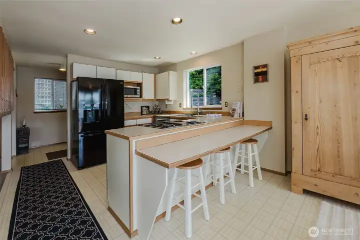 Bright, functional, and wide open — this kitchen is laid out the way a kitchen should be. The generous peninsula with warm wood-trimmed countertops does triple duty as prep space, cooktop station, and breakfast bar, with three stools tucked underneath for casual meals, morning coffee, or keeping the cook company while dinner comes together.  The cooktop sits right in the peninsula, keeping the chef in the center of the action and connected to the dining room and living spaces beyond — no one gets left out of the conversation. Recessed lighting overhead keeps the workspace bright and clean at any hour of the day.    The only thing missing is you!
