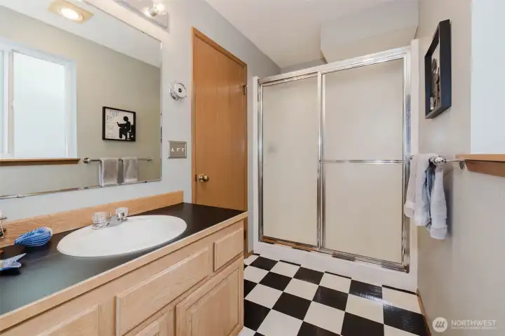 A bold black and white checkered floor sets a classic, timeless tone — the kind of design choice that feels intentional and fun rather than dated. It is the kind of detail that makes you smile the first time you see it and keeps making you smile every single day after.  Warm natural wood cabinetry carries through from the rest of the home, topped with a deep charcoal countertop and a generous vessel-style sink that feels both practical and polished. The large mirror above reflects light beautifully, keeping the space feeling open and bright. A full glass-enclosed shower with sliding doors completes the three-quarter bath — clean, classic, and completely move-in ready.