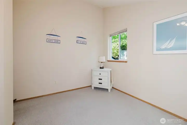 Bright, calm, and full of coastal charm — this second bedroom greets you with a smile. Cheerful "North Shore" and "At The Beach" signs hang on the wall, and while we will gently point out that this home actually sits on the beautiful South Shore of Hood Canal, the spirit is absolutely right. You are at the beach. You live at the beach. Every single day.  A garden-view window looks out to the lush greenery at the front of the home, filling the room with soft, peaceful natural light throughout the day. Plush neutral carpet, warm wood trim, and cream walls keep the space feeling clean, restful, and completely move-in ready.