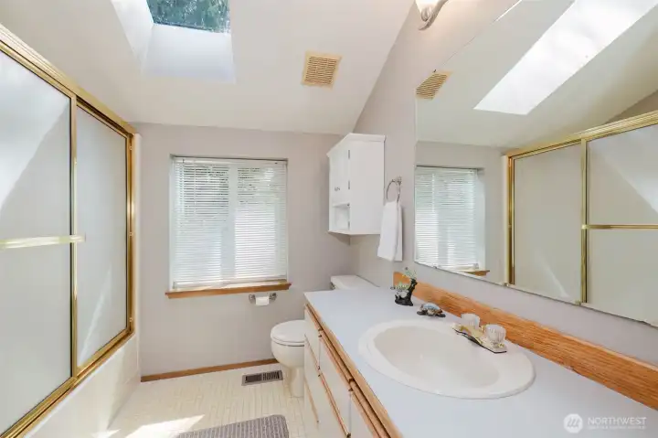 Natural light pours in from above at every hour of the day, giving this space an open, spa-like quality that you simply cannot fake with fixtures alone. On a sunny Pacific Northwest morning, this bathroom practically glows.  The vaulted ceiling adds unexpected height and airiness, while the large mirror spanning the full length of the vanity bounces that beautiful skylight illumination into every corner. Warm natural wood cabinetry with a freshly updated Soft Gray countertop feels clean, current, and move-in ready — a simple update that makes a genuine difference.  A full glass-enclosed shower with gold-toned sliding doors adds a classic, timeless touch, and the additional window brings in even more of that gorgeous green garden view from outside. White cabinetry above the toilet keeps everything organized and tidy, and the neutral tile floor and soft gray walls tie the whole space together quietly and cohesively.