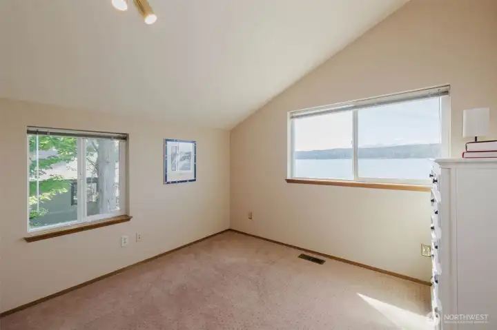 A generous waterfront-facing window frames a breathtaking panorama of Hood Canal and the mountains beyond, right from your bed. Before your feet even hit the floor, the water is there. Sparkling, shimmering, and entirely yours.  A second window on the adjacent wall looks out to the mature trees and greenery at the side of the home, wrapping the room in natural light from two directions and keeping it bright and airy from sunrise to sunset. The vaulted ceiling adds a sense of space and openness that makes this bedroom feel considerably larger than its footprint suggests.  Soft neutral carpet, warm cream walls, and track lighting overhead keep everything clean, calm, and completely move-in ready. There is nothing to do here but bring your furniture, hang your favorite art, and start living the waterfront life you have been dreaming about. Imagine the first morning you wake up here. Soft light on the water. The sound of the canal just outside. The mountains sitting quietly on the horizon.