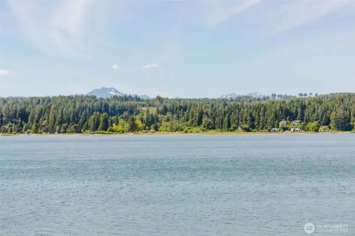 Snow-capped mountains rising behind a wall of Pacific Northwest evergreens, reflected in water so calm it barely seems real. This is what you see from your own deck, your own windows, your own life. No drive. No ticket. No waiting.  Some views you visit. This one you come home to.