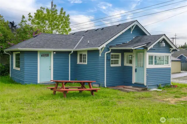This closer look highlights the charming blue siding, crisp white trim, and two separate entry doors—one into the cozy living room and another through the light-filled laundry/mudroom. The yard is flat and grassy with space for outdoor dining, gardening, or a potential patio upgrade, making this a versatile bonus for buyers looking for low-maintenance outdoor space.