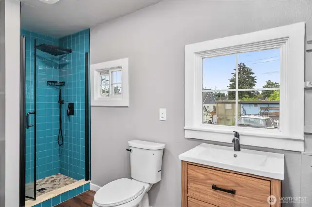 This modern bathroom combines eye-catching design with functional updates. A striking teal tile shower with black matte fixtures adds personality, while the large window and light wall tones keep the space feeling airy and bright. The sleek wood vanity brings warmth and style—proving that small spaces can still make a big impression.