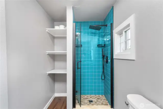 This bathroom makes the most of every inch, pairing a vibrant teal-tiled shower with sleek black fixtures and a rainfall showerhead. Built-in shelving provides convenient storage for linens and essentials—perfect for staying organized without sacrificing style.
