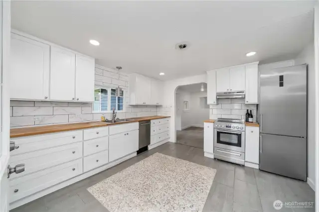 This light-filled kitchen features crisp white cabinetry, butcher block countertops, stainless steel appliances, and a stunning marble-style backsplash for a modern touch. A unique mosaic tile inlay adds character and defines the center of the space, while the open layout flows easily into the living area—ideal for entertaining or everyday comfort.