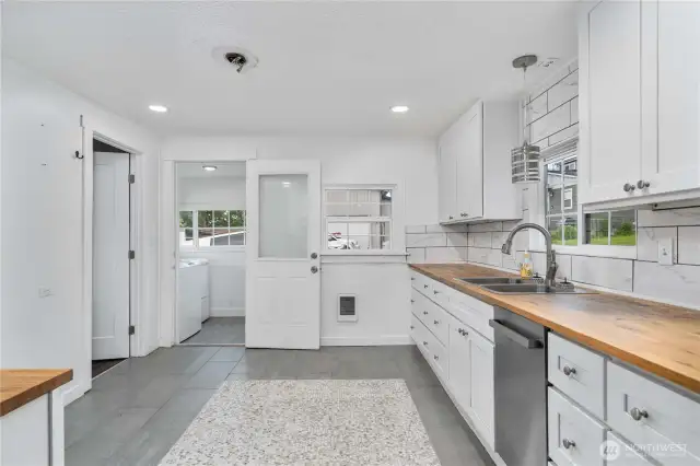 Enjoy a stylish, functional layout featuring butcher block countertops, sleek white cabinetry, and a beautiful tile backsplash. This view also highlights direct access to the bright mudroom/laundry area—perfect for keeping everyday messes out of sight and adding extra utility to this updated home.