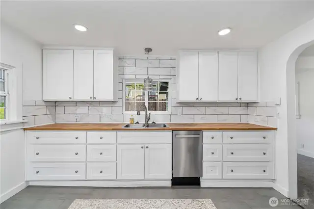 This crisp white kitchen blends charm and function with shaker-style cabinetry, warm butcher block counters, and a marble-look tile backsplash that adds a touch of elegance. A window above the sink brings in natural light, while the custom tile inlay underfoot defines the space with personality. Despite its compact footprint, there’s no shortage of necessities—including a sleek, space-saving dishwasher and plenty of cabinet storage for all your kitchen must-haves.