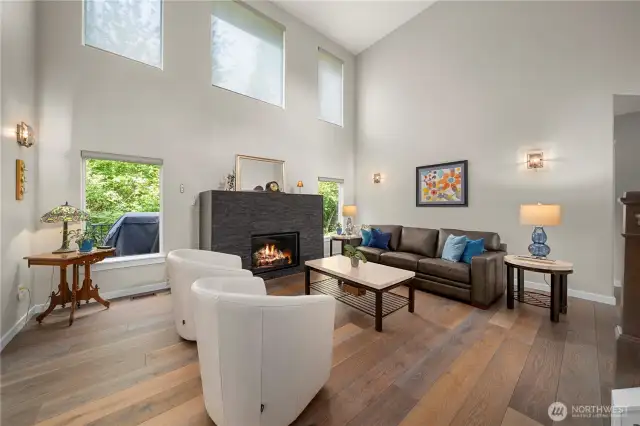 In the vaulted living room, custom silhouette blinds filter soft light through expansive windows, casting a glow across a bespoke limestone fireplace framed by a coffered ceiling.