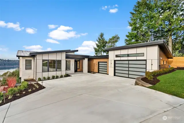 The striking exterior features a seamless blend of Hardie board siding, elegant stone accents, and frosted glass garage doors, perfectly complemented by a sleek black metal roof