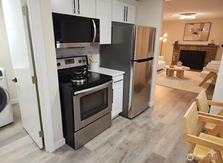 View from the kitchen toward the living room and laundry area — showcasing stainless steel appliances, modern finishes, and a seamless open flow between the main spaces.