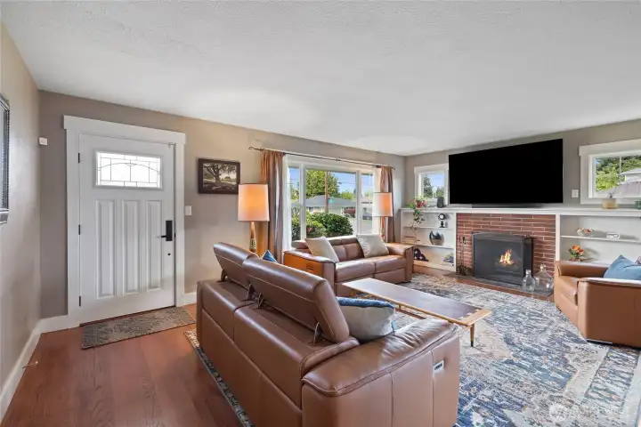 Light-filled living room featuring hardwood floors, oversized windows, custom built-ins, and a brick fireplace at center. Large picture window in front and side windows grace the fireplace with built in shelves.