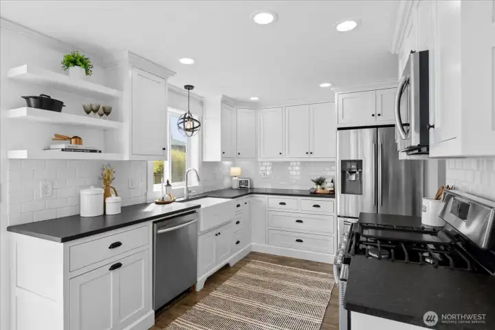 This modern kitchen is fully equipped with custom white cabinetry, quartz countertops, a large farmhouse sink, SS appliances, and tile backsplash.