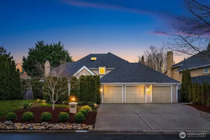Stunning twilight curb appeal showcases this impeccable Buchan home in the coveted Trossachs community, featuring a three-car garage, mature landscaping, and warm exterior lighting set against a dramatic Pacific Northwest sunset sky.