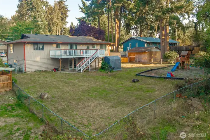 View from the back fence highlights the home’s full deck, lower patio, and large, fully fenced yard — ideal for pets, play, or future landscaping ideas. Mature trees and established greenery add a peaceful Pacific Northwest feel.