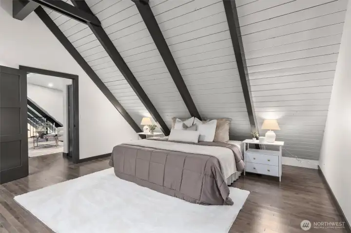 Welcome to a primary suite that feels like a boutique luxury hotel. Architecturally spectacular, this grand sanctuary boasts soaring, sharply angled cathedral ceilings lined with clean white planking and anchored by bold dark beams. Geometric accent windows frame peaceful views of the surrounding treetops, filling the room with soft, natural light.  ?The luxury continues with a massive his-and-hers walk-in closet fully equipped with custom built-in shelving to keep your wardrobe flawlessly organized.