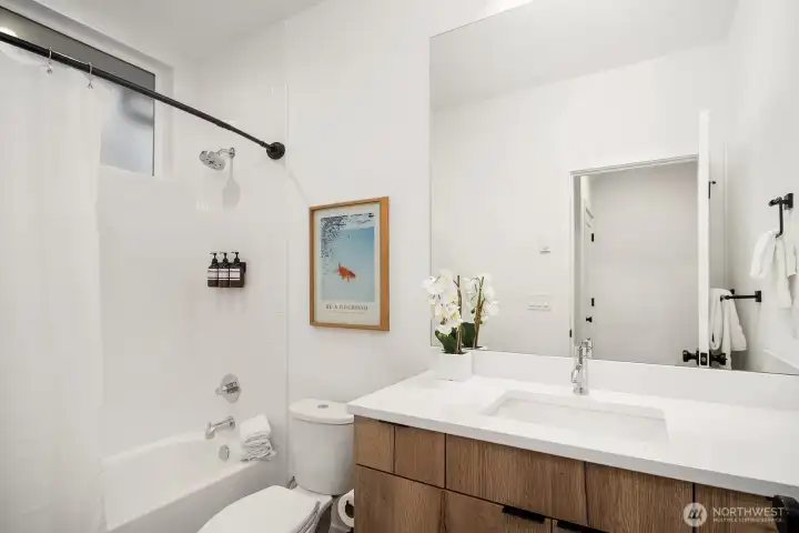 Clean and well-appointed full guest bath with a warm wood vanity, tiled tub and shower combo, and modern fixtures delivering a polished finish on the entry level.