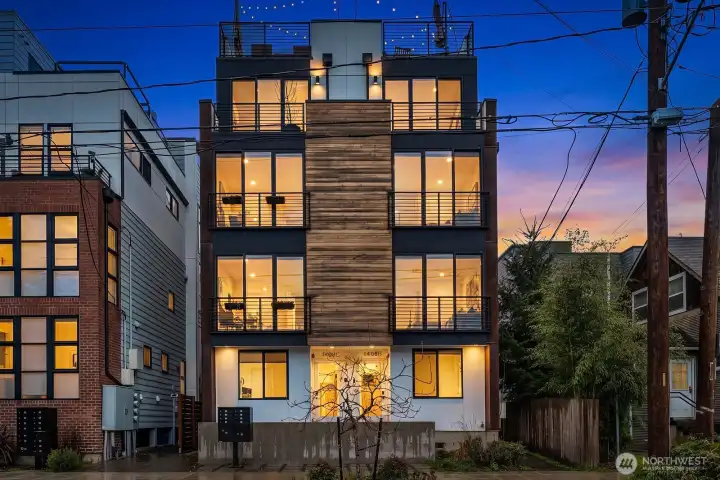 This striking modern end-unit townhome glows at dusk, showcasing its contemporary architecture and floor-to-ceiling windows that hint at the light-filled interiors within.