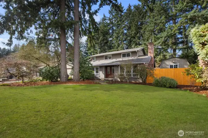 Expansive front yard with manicured lawn, mature evergreen trees, and landscaped garden beds leading to the covered front entry of this two-story Puyallup home.