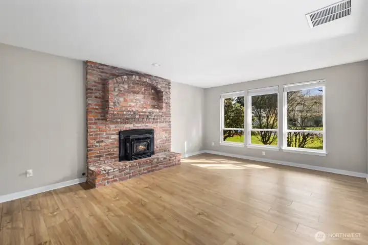 Bright living room with large front-facing windows, wood-style flooring, and a brick fireplace with insert creating a warm focal point.