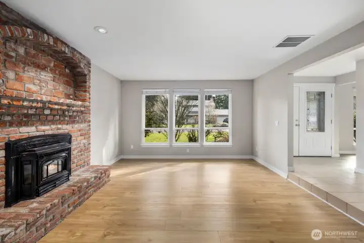 Open living room with natural light from large windows, brick fireplace feature wall, and direct flow toward the entry and main living areas.