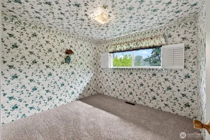 Charming bedroom with vintage-style floral walls creates a cozy cottage feel, complemented by plush carpeting and a bright window that frames treetop and garden views. This versatile room works beautifully as a nursery, office, or guest space depending on your needs.