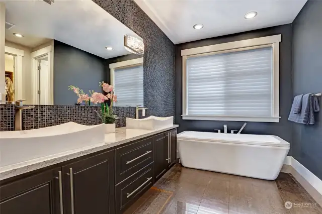 Primary spa-like bathroom with elevated cabinets, under cabinet lighting, double vessel sinks, linen cabinet to the left of the left sink, full-length mosaic tile accent wall, and a tub fit for relaxation and bubble baths.