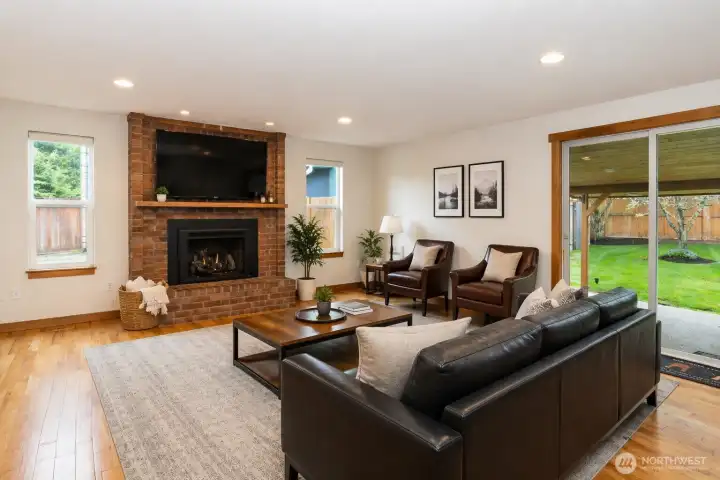 "Virtually Stage" Spacious rec room featuring a cozy brick fireplace, hardwood floors, and recessed lighting. Large sliding glass door opens to a covered patio and fully fenced backyard, creating seamless indoor-outdoor living—ideal for entertaining, or relaxation space.