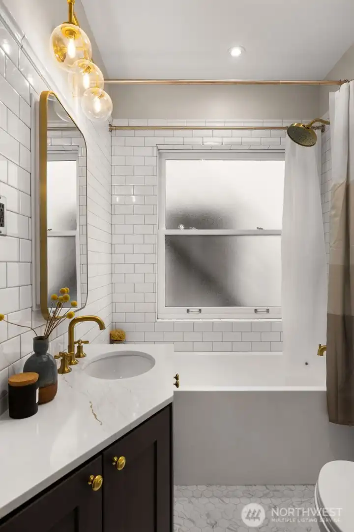 The bathroom offers a deep soaking tub surrounded by subway-tiled walls, elegant brass fixtures & hexagon ceramic tile floors.