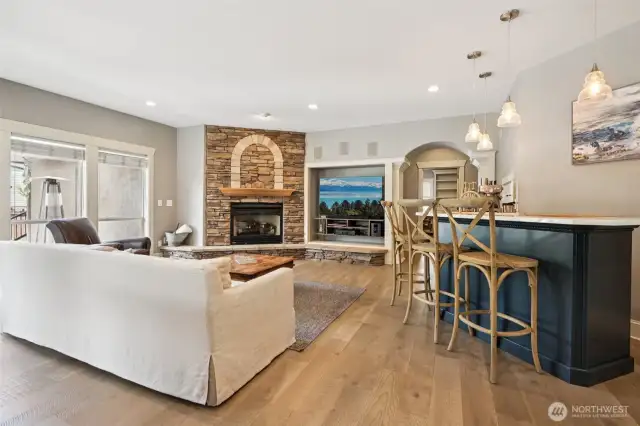A seamless open-concept living space designed for effortless entertaining. The grand stone fireplace anchors the room with style, while natural light pours in from oversized windows. The expansive kitchen bar invites casual conversations and cozy evenings in, all set against wide-plank oak floors and rich architectural detail that elevates every moment