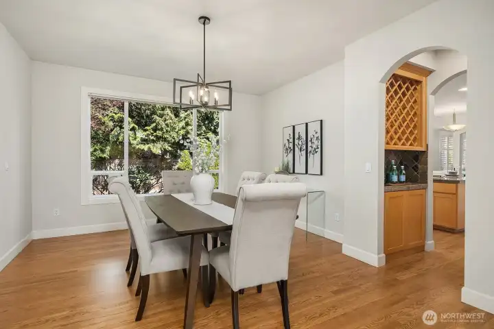 This alternate view of the dining room further highlights the bright, airy atmosphere created by the large garden-facing window, elegant chandelier, and the warm hardwood floors that flow seamlessly into the adjacent living spaces. The graceful arched doorway leading to the kitchen beyond adds a sophisticated architectural detail that elevates the home's overall sense of refinement.