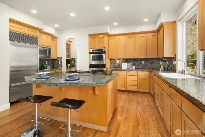This inviting view of the kitchen showcases the central island with bar seating as the heart of the space, perfectly positioned for casual meals, homework, or entertaining while the chef prepares dinner. The warm wood cabinetry, granite and slate counters, stainless steel appliances, and recessed lighting come together to create a kitchen that is both highly functional and timelessly beautiful.