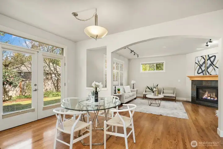 This bright and inviting space seamlessly blends a casual dining nook and family room, bathed in abundant natural light from a wall of windows that frame beautiful views of the lush surroundings. The cozy fireplace, rich hardwood floors, and vaulted ceilings create a warm and relaxed atmosphere perfect for everyday family living and informal gatherings.
