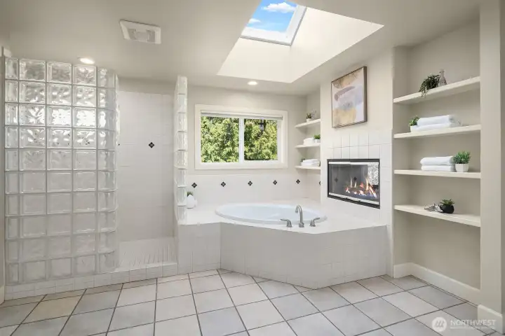 This indulgent corner of the primary bath features a luxurious soaking tub bathed in natural light from a skylight above, perfectly positioned beside a glass block window and elegant built-in shelving for a serene, spa-like atmosphere. Every detail of this beautifully designed space invites relaxation and reflects the exceptional level of comfort and refinement found throughout this remarkable home.