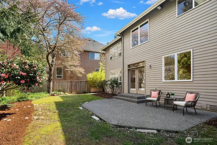 This alternate view of the backyard showcases the seamless transition from the home's rear exterior to the inviting patio and lush, fully fenced lawn beyond, highlighting the generous outdoor living space available for relaxing and entertaining. The mature surrounding landscaping, full irrigation system, and private setting make this backyard a true extension of the home's exceptional living experience.