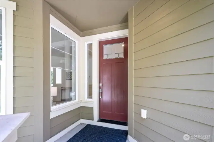 A deep chestnut paneled front door adds friendly color beneath the sheltered porch, with a leaded-glass-style window for character and light. The satin-nickel entry handleset pairs nicely with the Craftsman-inspired exterior, while nearby windows brighten the front entry.