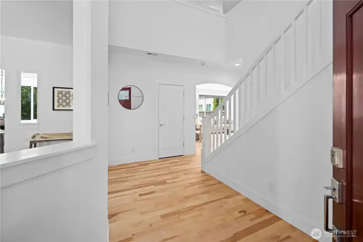 Step inside to a grand cathedral-ceiling foyer, where warm hardwood floors, fresh white millwork, and an open staircase set an elegant first impression. A formal living room opens to the left, while large windows, high ceilings, and the arched opening ahead give the entry a bright, open sense of arrival.