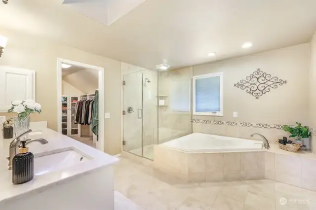 Primary bath features natural earth-tone travertine tile flooring, a double vanity with quartz countertop, large soaking tub, and travertine tile walk-in shower wrapped with seamless glass surround.