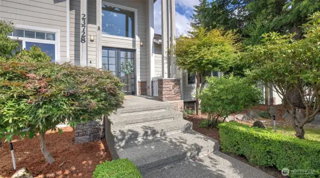 Grand entryway framed by lush greenery, featuring elegant double glass doors and a striking transom window above!