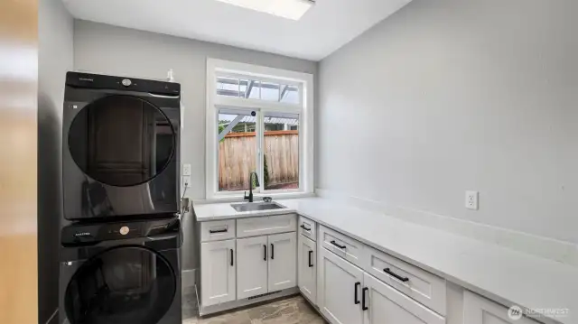 A generously sized laundry area featuring crisp white cabinetry with black handles, ample storage, and a built-in sink for added functionality