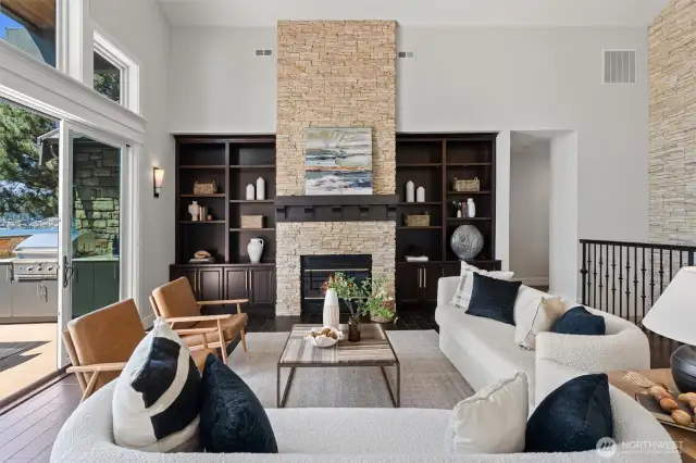 A vaulted great room with a wall of windows and built-in cabinetry surrounds a stunning stone fireplace, all overlooking sweeping lake views and a spacious deck.